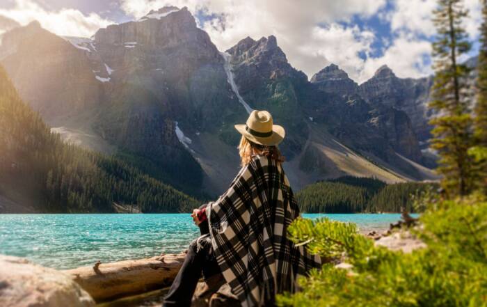 Une personne portant un chapeau et un plaid à carreaux contemple de dos un lac turquoise bordé de sapins et d'imposantes montagnes rocheuses ensoleillées.