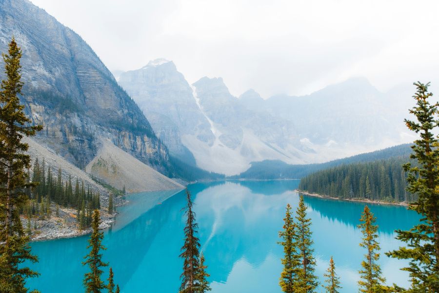 Un lac turquoise étincelant reflète des montagnes enneigées et escarpées, bordé de pins verts sous un ciel brumeux.