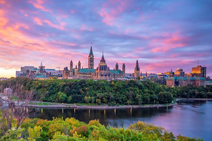 Les édifices historiques du Parlement surplombent une colline verdoyante et une rivière, sous un ciel aux teintes roses et violettes.