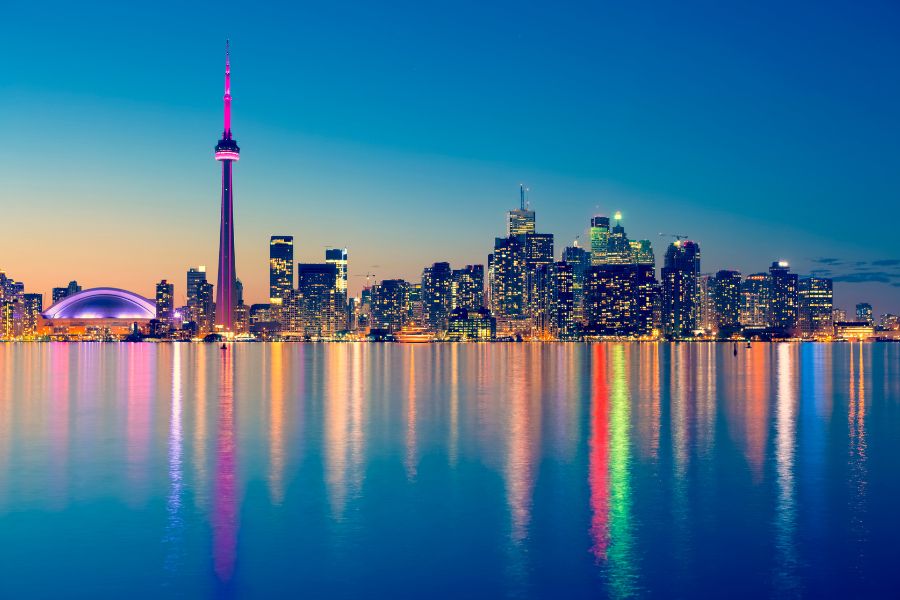 Un panorama urbain s'illumine au crépuscule, projetant des reflets colorés et allongés sur les eaux calmes au premier plan.