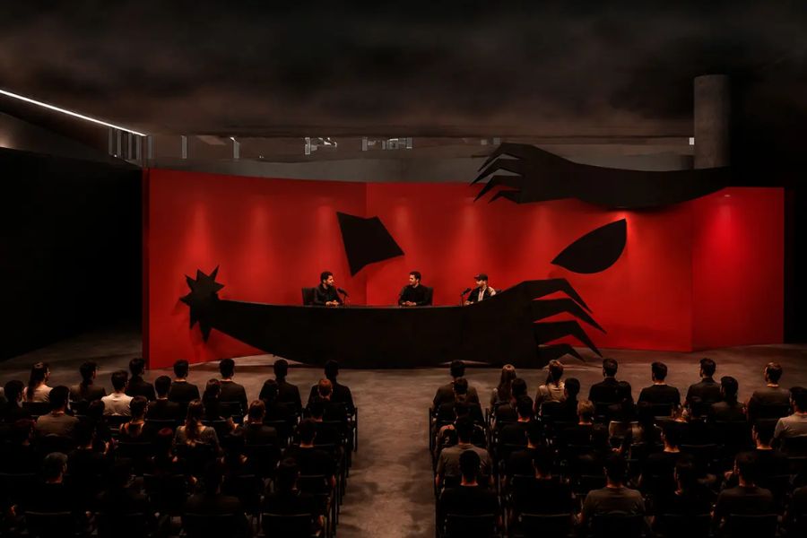 Trois personnes siègent derrière une table sombre sur une scène encadrée d'un décor rouge et noir en forme de monstre, face à un public attentif assis dans une salle obscure.