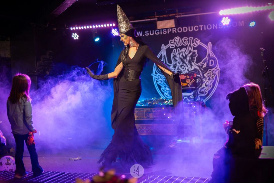 Une personne déguisée en sorcière avec un chapeau pointu effectue une performance sur une scène enfumée et éclairée de violet, sous l'inscription « W.SUGISPRODUCTIONS.COM », devant un jeune public.