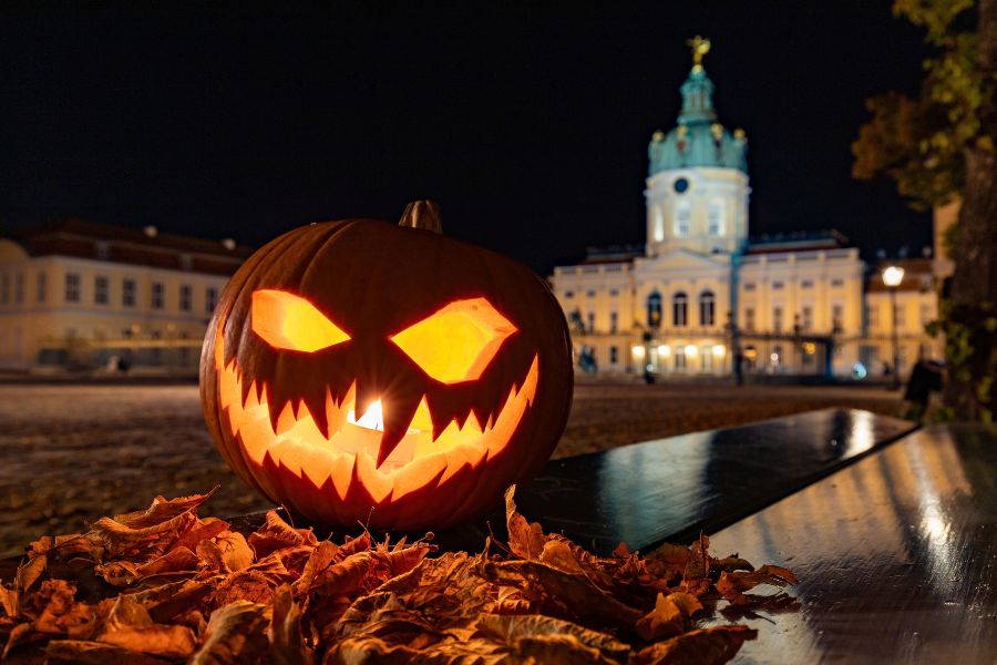 Une citrouille illuminée au visage effrayant repose sur un lit de feuilles mortes au premier plan, devant un château éclairé en arrière-plan la nuit.