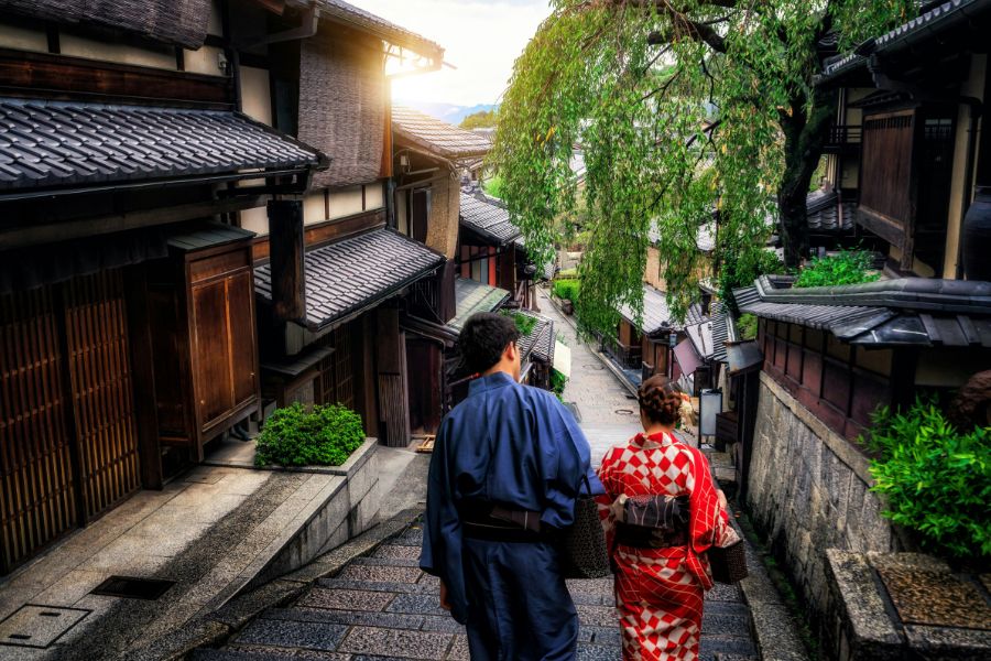 Un homme et une femme vêtus de kimonos traditionnels descendent une ruelle pavée en pente, bordée de maisons anciennes en bois aux toits de tuiles.