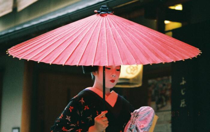 Une geisha vêtue d'un kimono sombre tient une ombrelle traditionnelle en papier rouge tout en marchant dans une rue devant un bâtiment japonais.