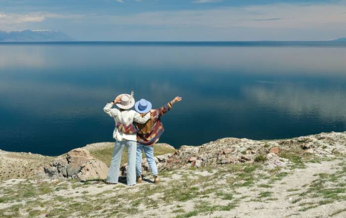 Deux personnes en tenues légères et chapeaux s'enlacent de dos au sommet d'une falaise herbeuse, désignant l'horizon au-dessus d'une vaste étendue d'eau calme.