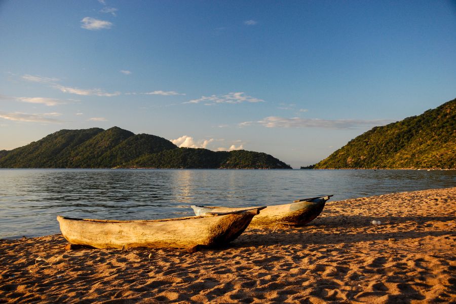 Deux pirogues en bois reposent sur une plage de sable fin au bord d'un lac bordé de montagnes verdoyantes.