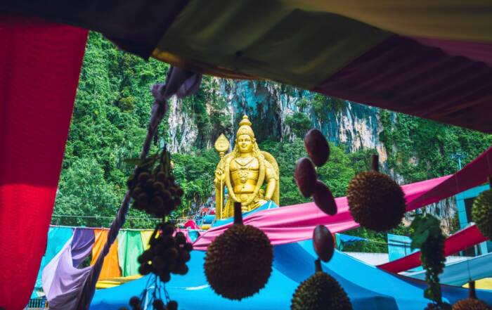 Une grande statue dorée du dieu Murugan se dresse devant une falaise verdoyante, cadrée par des tentures colorées et des fruits suspendus.