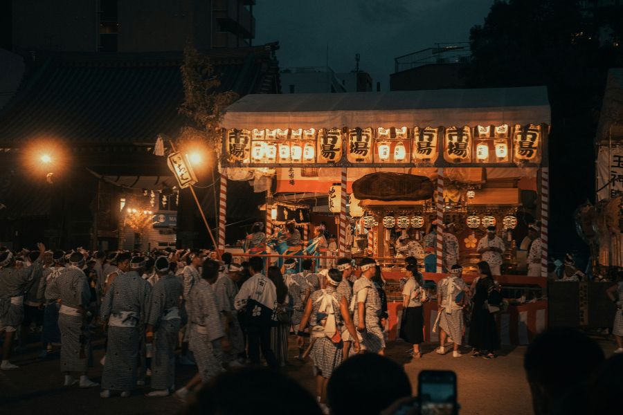 Une foule vêtue d'habits traditionnels défile de nuit devant un stand illuminé de nombreuses lanternes en papier suspendues sous une structure décorée.