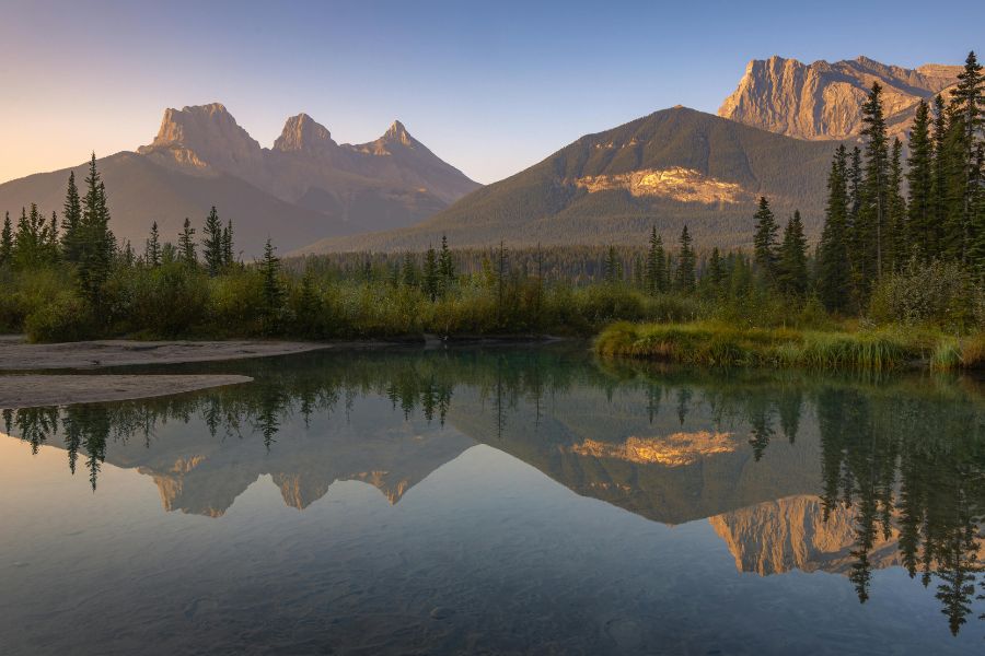 De grands sommets rocheux se reflètent nettement sur l'eau calme d'une rivière bordée de conifères sous une douce lumière matinale.