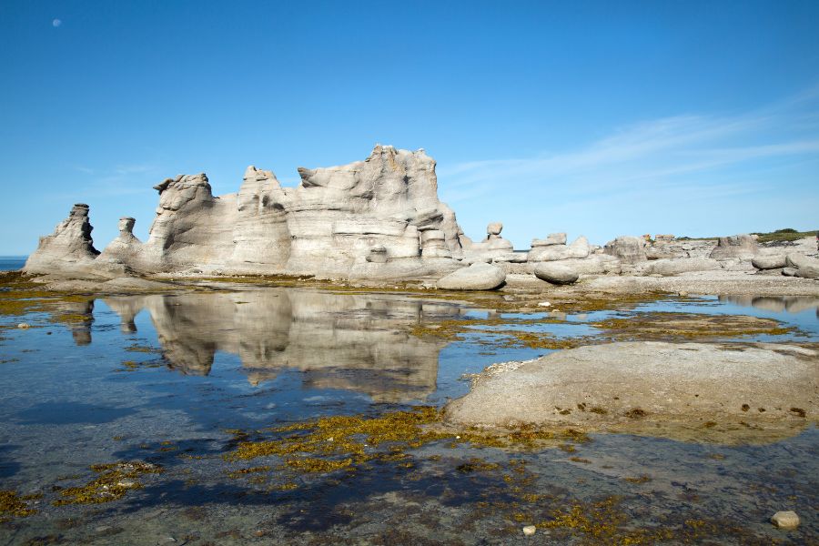 Des monolithes de pierre calcaire sculptés par l'érosion se dressent sur un rivage rocheux bordé de mares d'eau sous un ciel bleu.