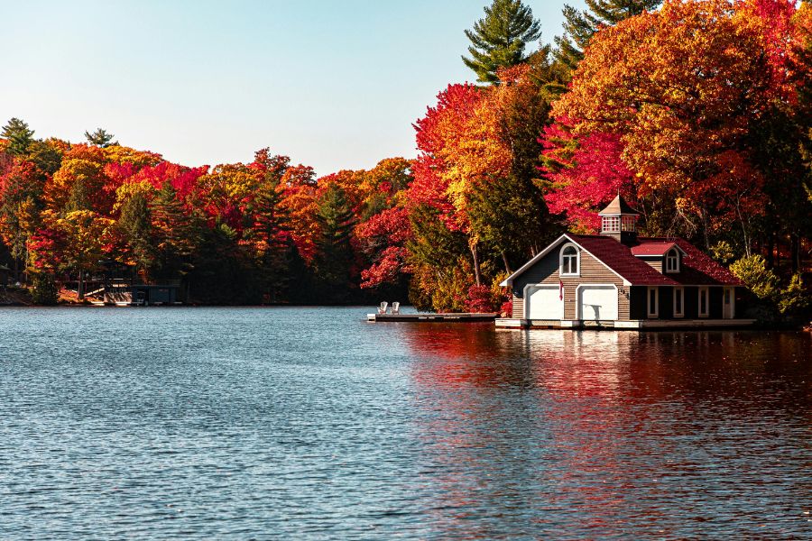 Un garage à bateaux en bois borde un lac entouré d'une forêt aux feuillages d'automne rougeoiants et dorés.