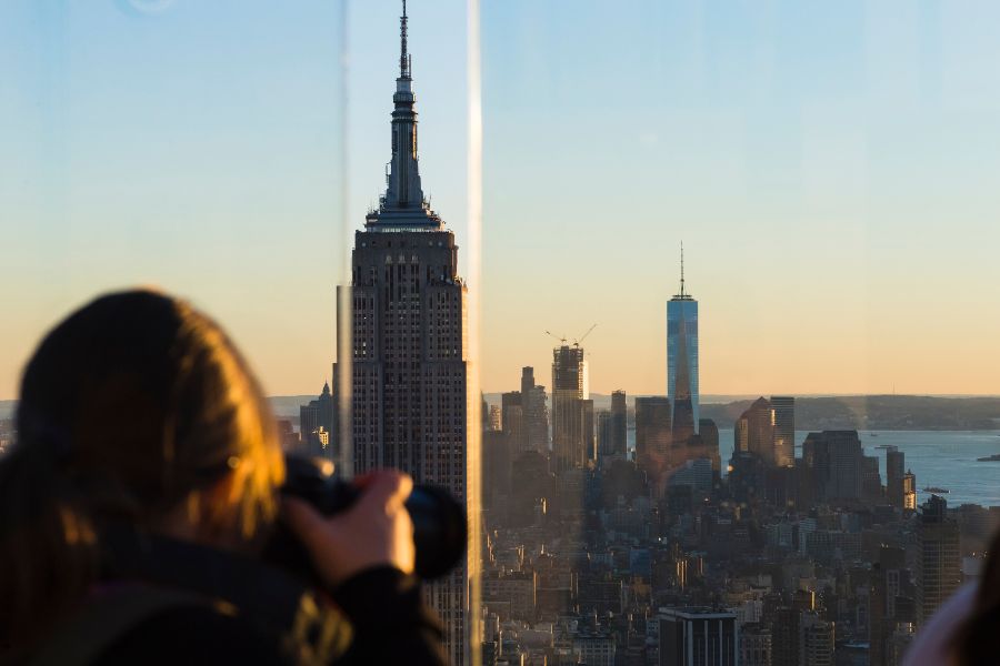 Une personne prend en photo à travers une baie vitrée les célèbres gratte-ciel d'une métropole s'étendant sous la lumière dorée du soleil couchant.