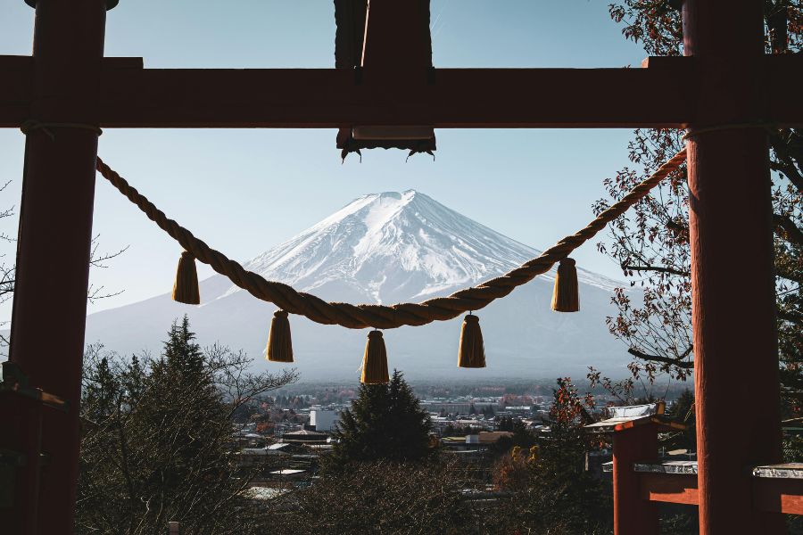 Une grosse corde sacrée suspendue à la structure en bois d'un portail torii encadre le sommet enneigé du mont Fuji se découpant sous un ciel clair.