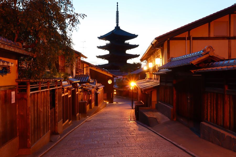 Une pagode en bois à plusieurs étages se dresse au bout d'une ruelle pavée bordée de maisons traditionnelles éclairées par des lanternes au crépuscule.