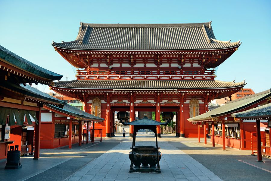Une grande porte monumentale en bois rouge et toit de tuiles domine une cour pavée abritant un brûleur d'encens central devant des pavillons.