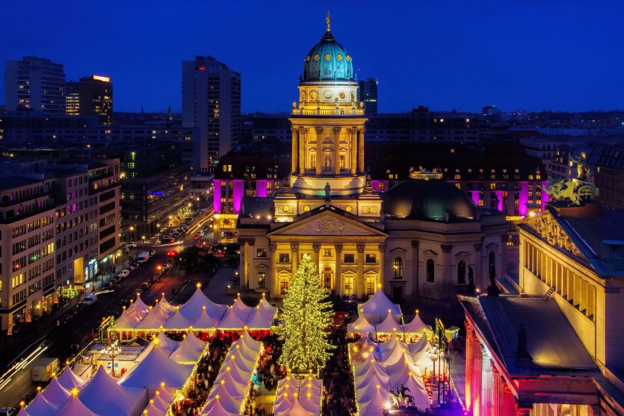 Des tentes de marché illuminées entourent un grand sapin de Noël scintillant au pied d'une église historique éclairée la nuit.