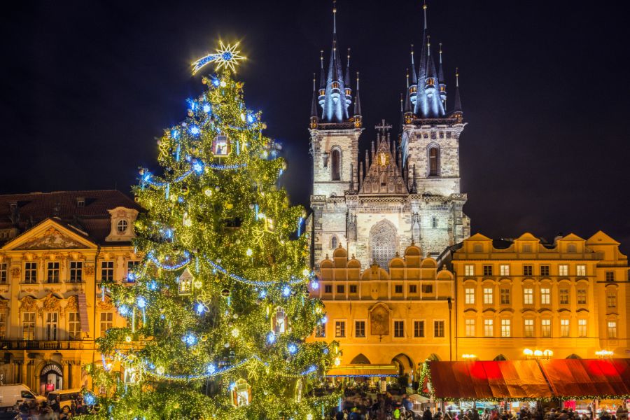 Un grand sapin de Noël décoré de lumières dorées brille sur une place pavée dominée par les tours éclairées d'une église gothique.