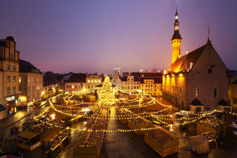 Des guirlandes lumineuses rayonnent depuis un sapin central vers les chalets en bois d'une place médiévale dominée par un beffroi.