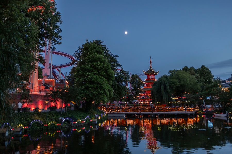 Des lanternes et des attractions illuminées se reflètent sur un lac paisible bordé d'arbres et d'une pagode traditionnelle au crépuscule.