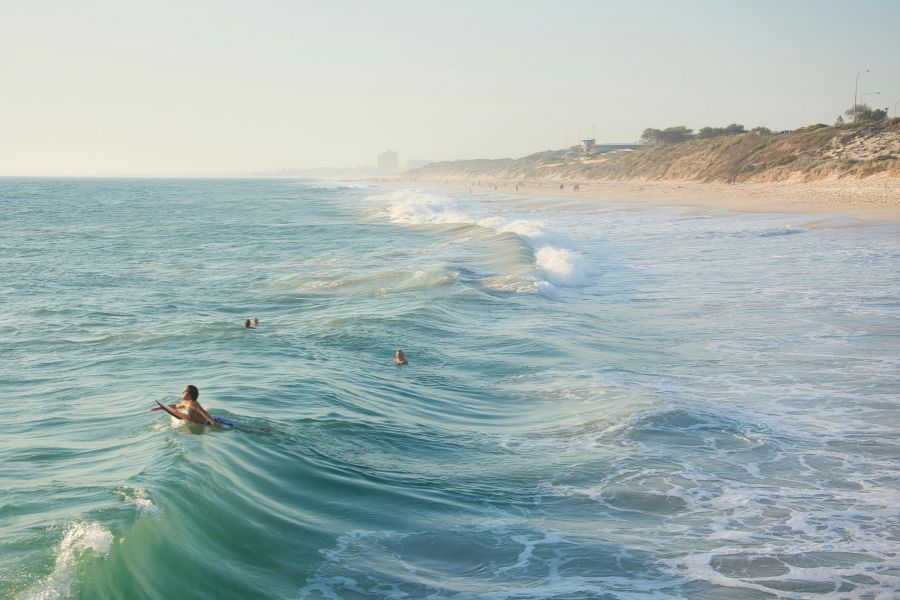 Des baigneurs profitent des vagues dans une eau claire le long d'une vaste plage de sable bordée de dunes verdoyantes.