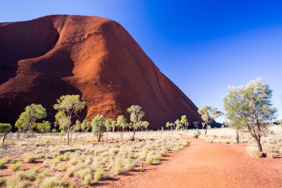 Un chemin de terre rouge serpente à travers une végétation aride au pied d'un immense monolithe rocheux sous un ciel bleu intense.