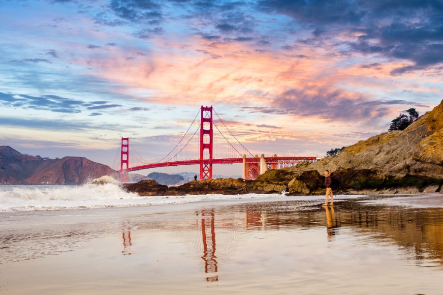 Une personne se tient sur une plage de sable humide reflétant les couleurs d'un coucher de soleil flamboyant derrière un grand pont suspendu rouge.